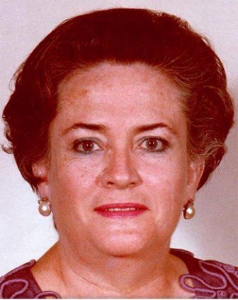 Woman with brown hair, wearing pearl earrings, looking forward, red lipstick, and a patterned top.
