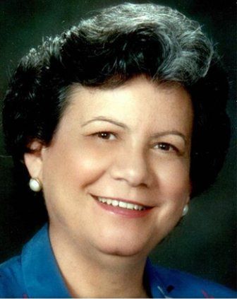 Woman with dark, curly hair and pearl earrings smiles; wearing blue top.