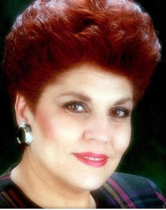 Woman with auburn hair, wearing black earrings and a patterned top, smiles.