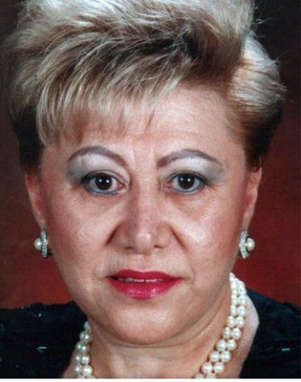 Woman wearing pearl necklace and earrings, looking at the camera. She has blonde hair and red lipstick.