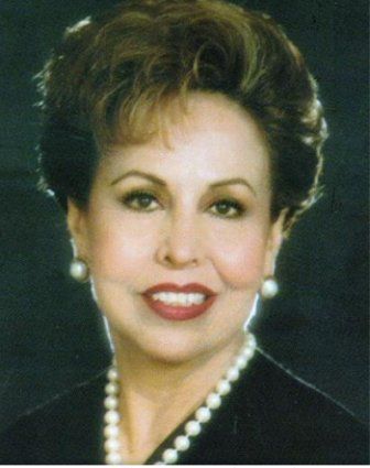 Woman with short, curly brown hair, wearing a pearl necklace and earrings, smiling.