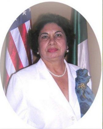 Woman in white blazer stands before US and Mexican flags, smiling.