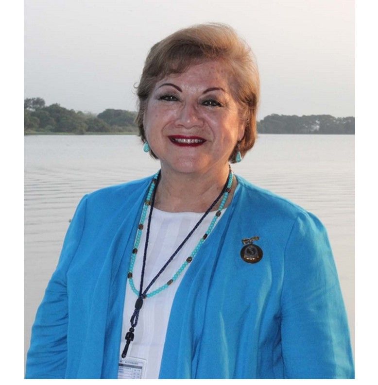 Woman in turquoise jacket and beaded necklace, smiling in front of water.