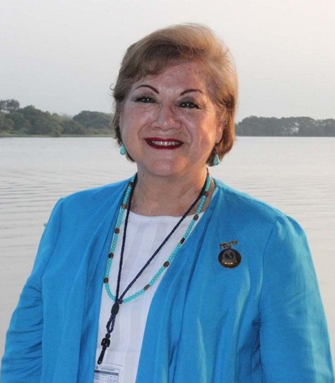 Woman in blue blazer, smiling, standing near a body of water; wears turquoise jewelry, a pin on her blazer.