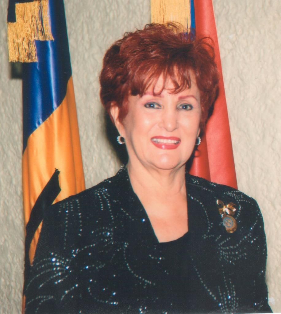 Woman with short red hair in a black sequined jacket smiles. Flags in the background.