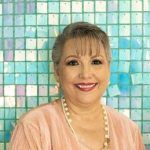 Woman smiling, wearing a pink blouse and pearl necklace, in front of a blue and white tiled wall.