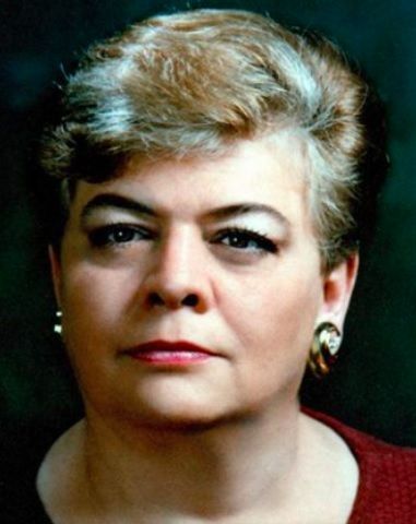 Woman with short blonde hair and gold earrings, wearing a red top, looking directly at the viewer.