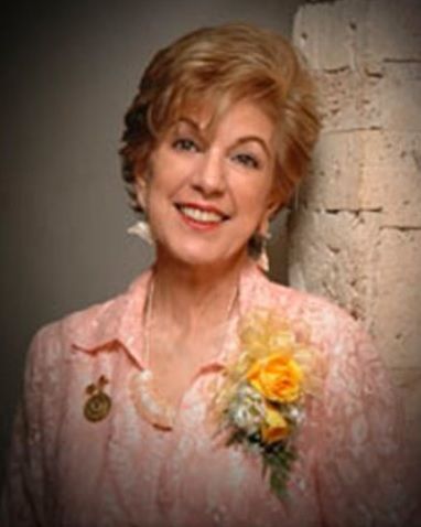 Woman with short, light hair wearing a pink blouse and smiling, with a flower pin.