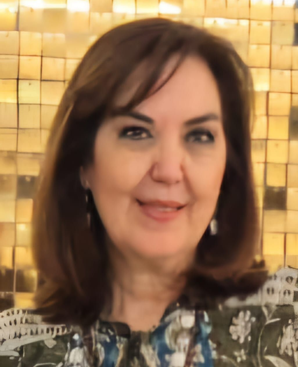 Woman smiling in front of a textured gold background. She has brown hair and is wearing a patterned top.