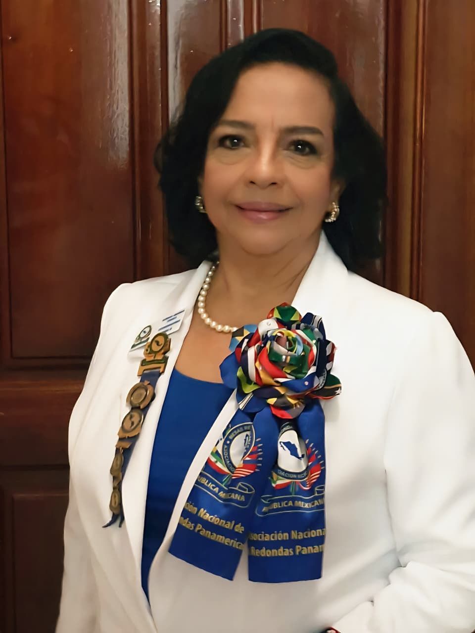Woman in a white jacket, blue top, and pearl necklace, wearing a rosette. Standing in front of a wooden wall.