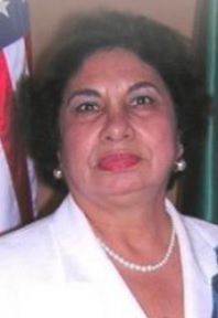 Woman wearing a white blazer and pearl necklace, smiling; flags in the background.