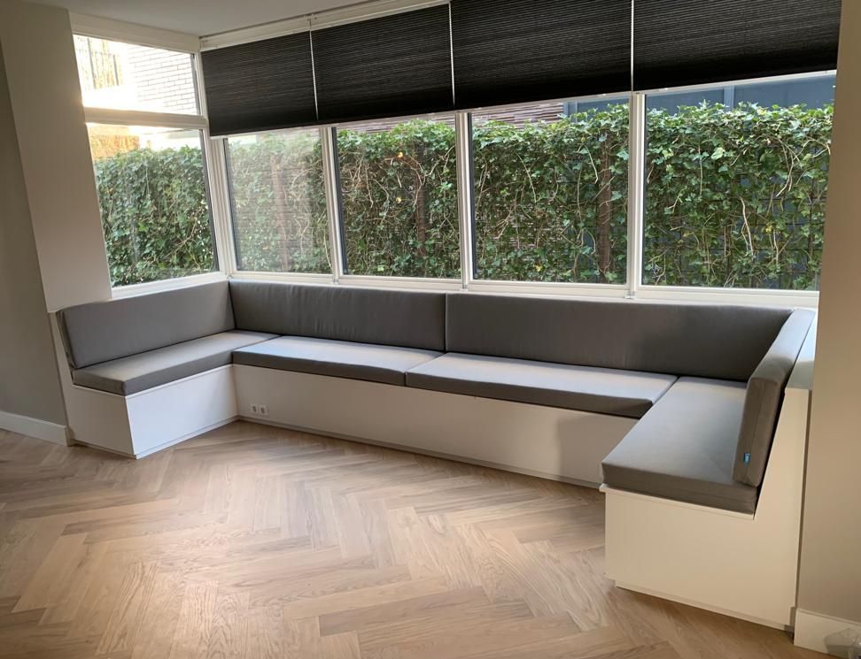 Ingebouwde witte maatwerk zitbank met grijze kussens naast een groot raam met uitzicht op groen. Erker. Interieuradvies.