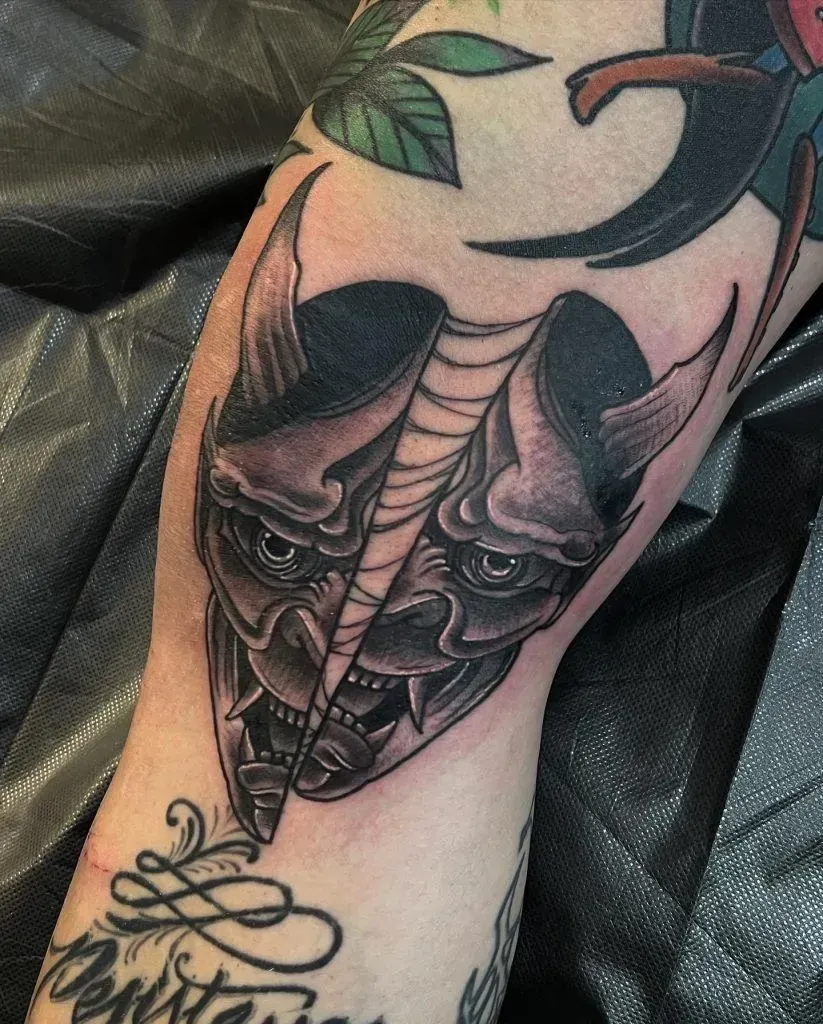 Tattoo of a split, horned demon mask on a person's skin. Gray scale with black shading and detail.