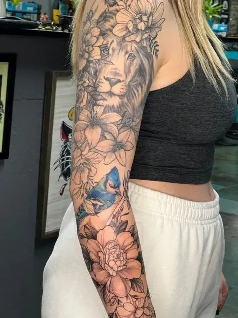 Tattoo of a lion's face and flowers on a person's arm, with a bluebird and a gray crop top.