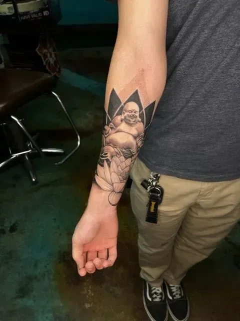 Tattoo on arm depicting a laughing Buddha figure within a lotus-like design. The person wears a gray shirt and khaki pants.