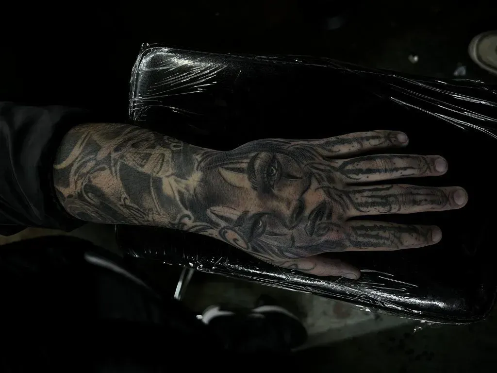 Tattooed hand in black substance, possibly liquid. Arm visible. Dark setting.
