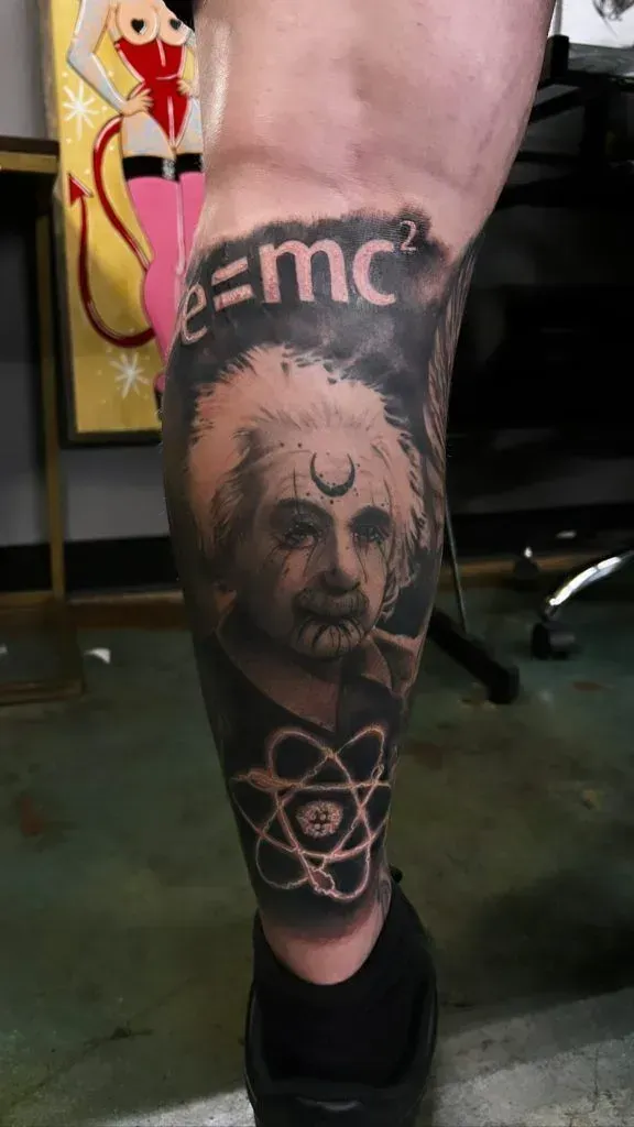 Tattoo of Einstein with formula, atom, and crescent moon on a leg. Black and gray, indoors.