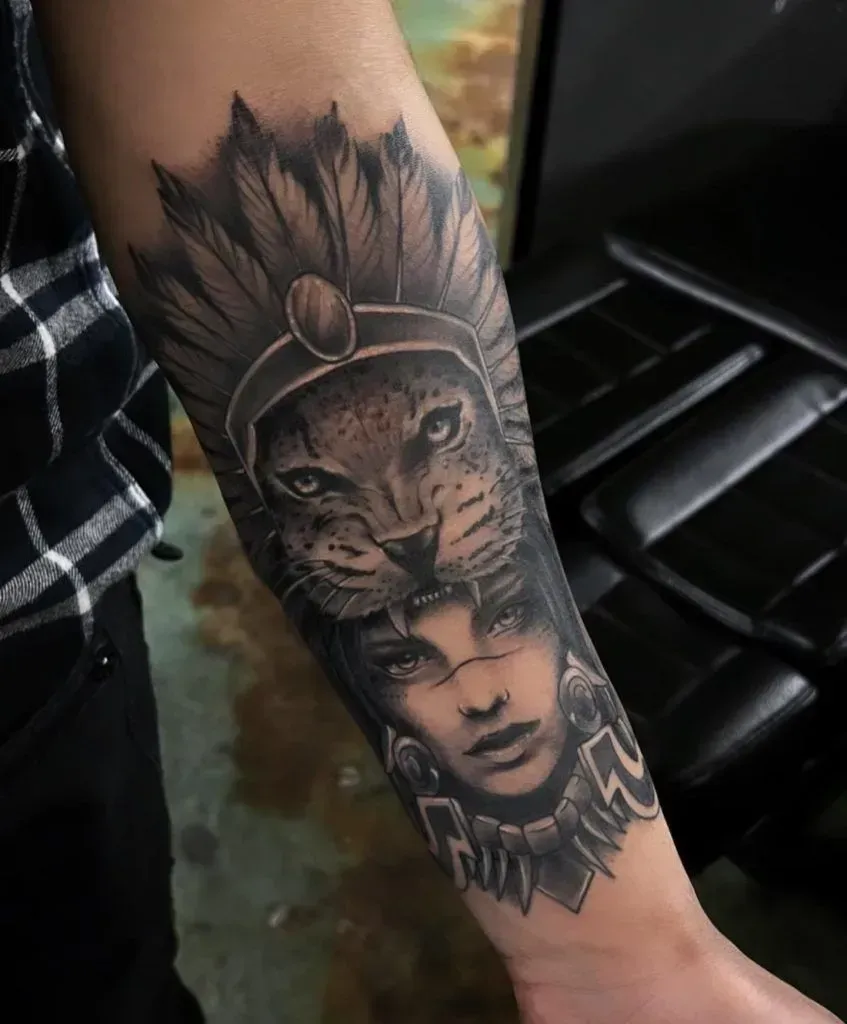 Tattoo of a panther head over a woman's face, wearing a feathered headdress. Arm in black and gray.