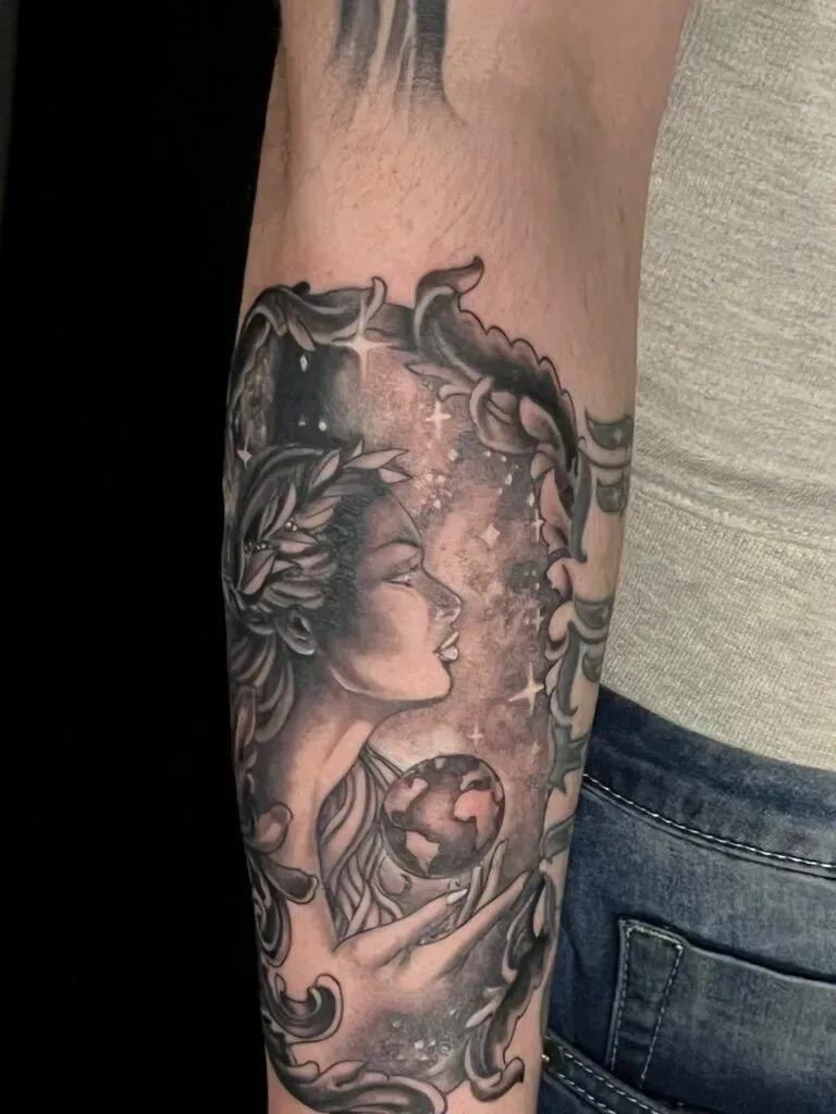 Tattoo of a woman with laurel wreath profile, holding a globe, surrounded by ornate frame and stars.