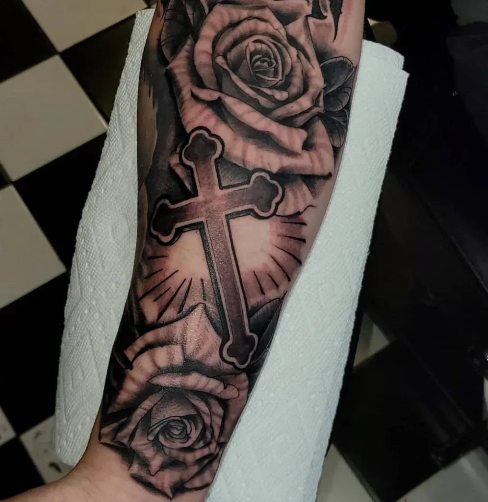 Tattoo of a cross with rays of light, surrounded by roses on an arm, black and white checkered background.