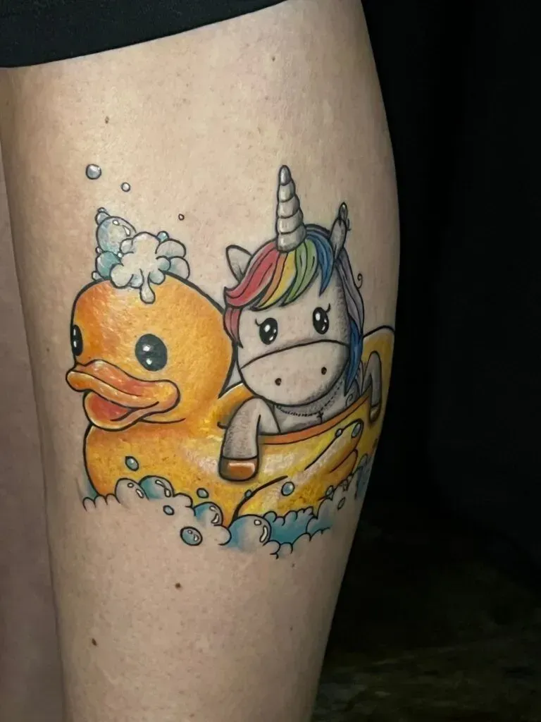 Tattoo of a unicorn in a rubber duck inner tube. The duck is yellow, the unicorn has a rainbow mane, with bubbles.