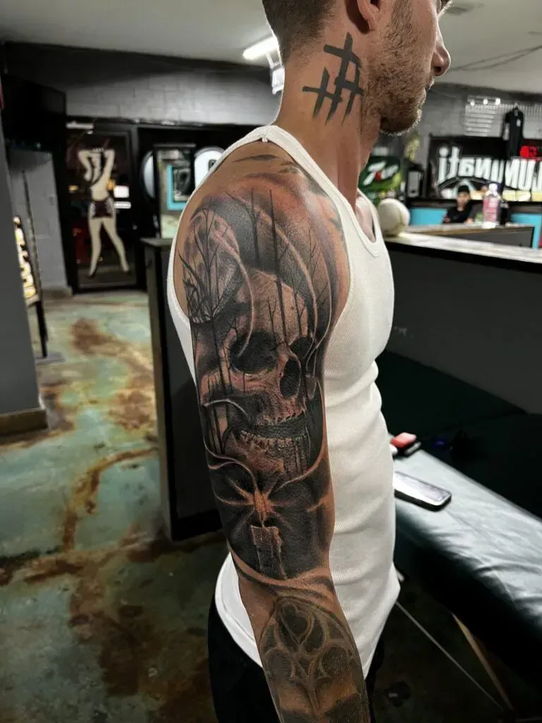 Man with a detailed black and grey skull sleeve tattoo. He's wearing a white tank top, in a tattoo parlor setting.