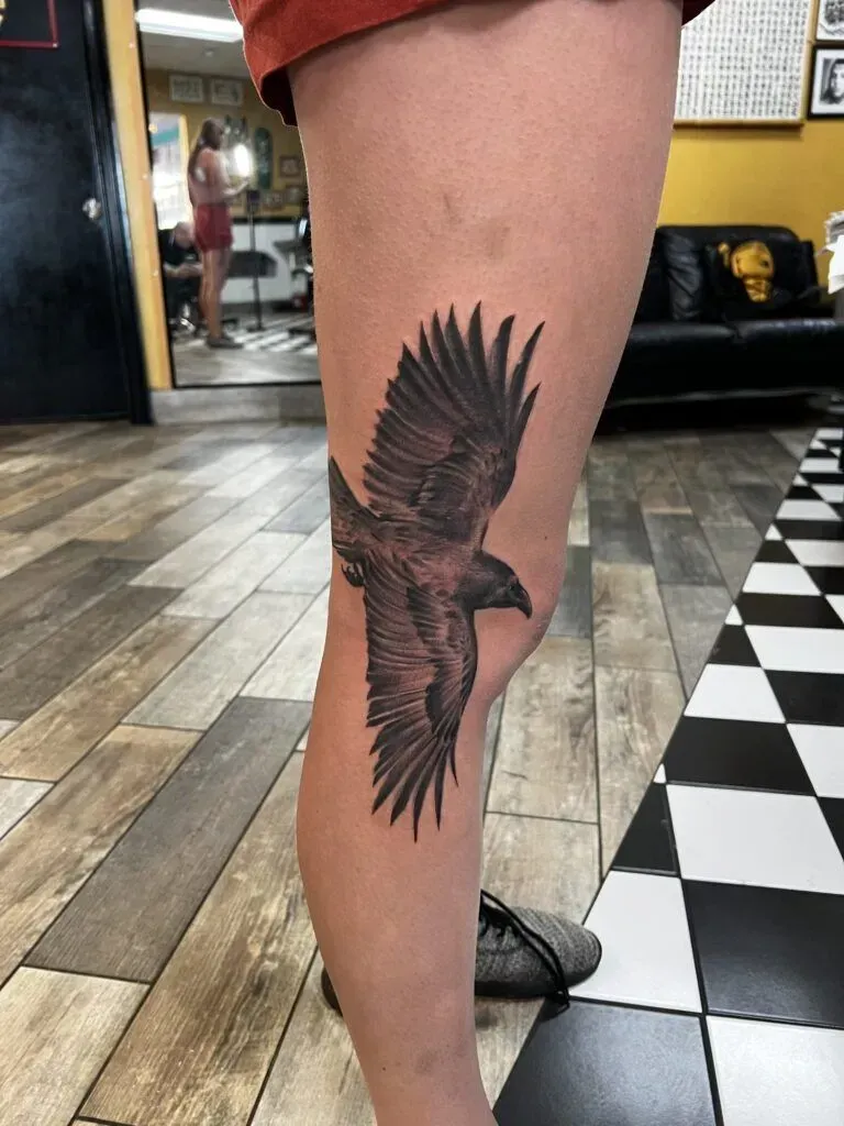 Tattoo of a dark eagle with spread wings on a person's leg. In a tattoo parlor setting.
