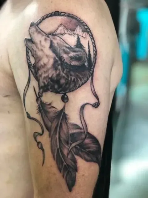 Tattoo of a howling wolf inside a dreamcatcher, with feathers dangling below, on an upper arm.