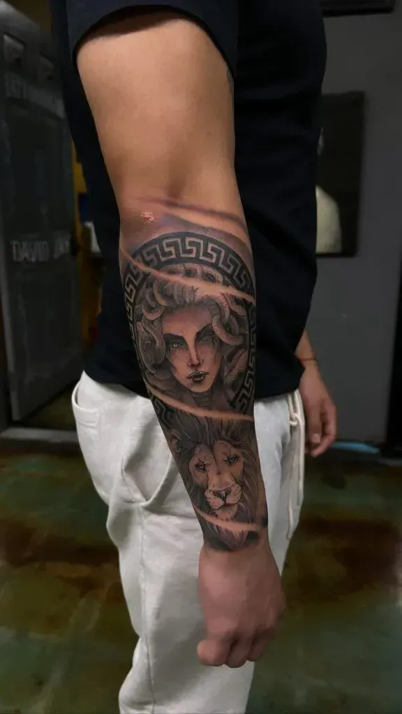 Man's forearm tattoo featuring a Medusa head and lion face with ornate Greek key border in grayscale.