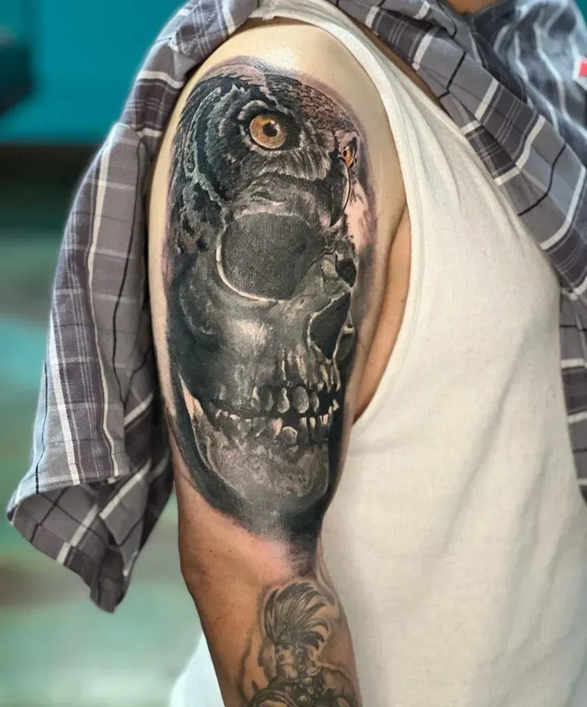 Tattoo of a skull and owl eye on a person's upper arm, in grayscale. A plaid shirt is draped on the shoulder.