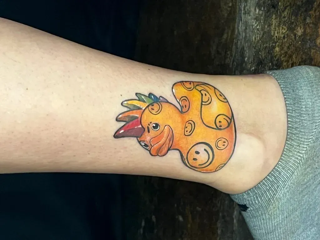 Cartoon character tattoo on ankle with orange body, smiley faces, and rainbow feathers.