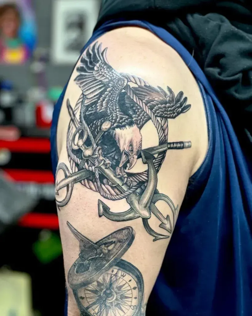 Tattoo of an eagle gripping an anchor with a nautical compass below on a person's upper arm.