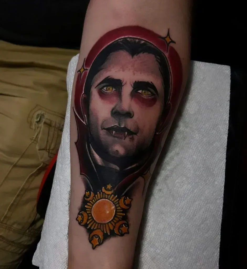 Tattoo of a vampire with red eyes on a forearm, within a red frame and a golden pendant at the bottom.