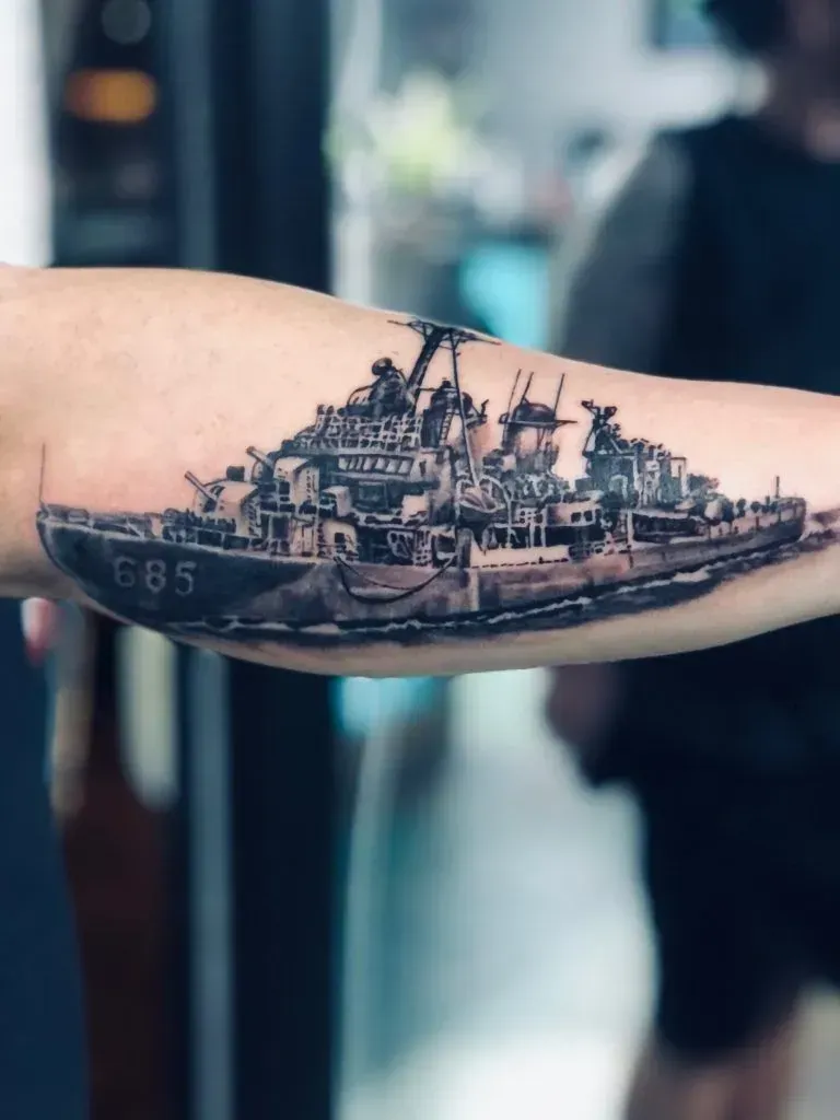 Tattoo of a battleship on a forearm in black and gray ink;