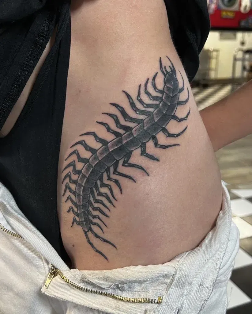 Black and gray centipede tattoo on a person's side, against a black and white tiled floor.