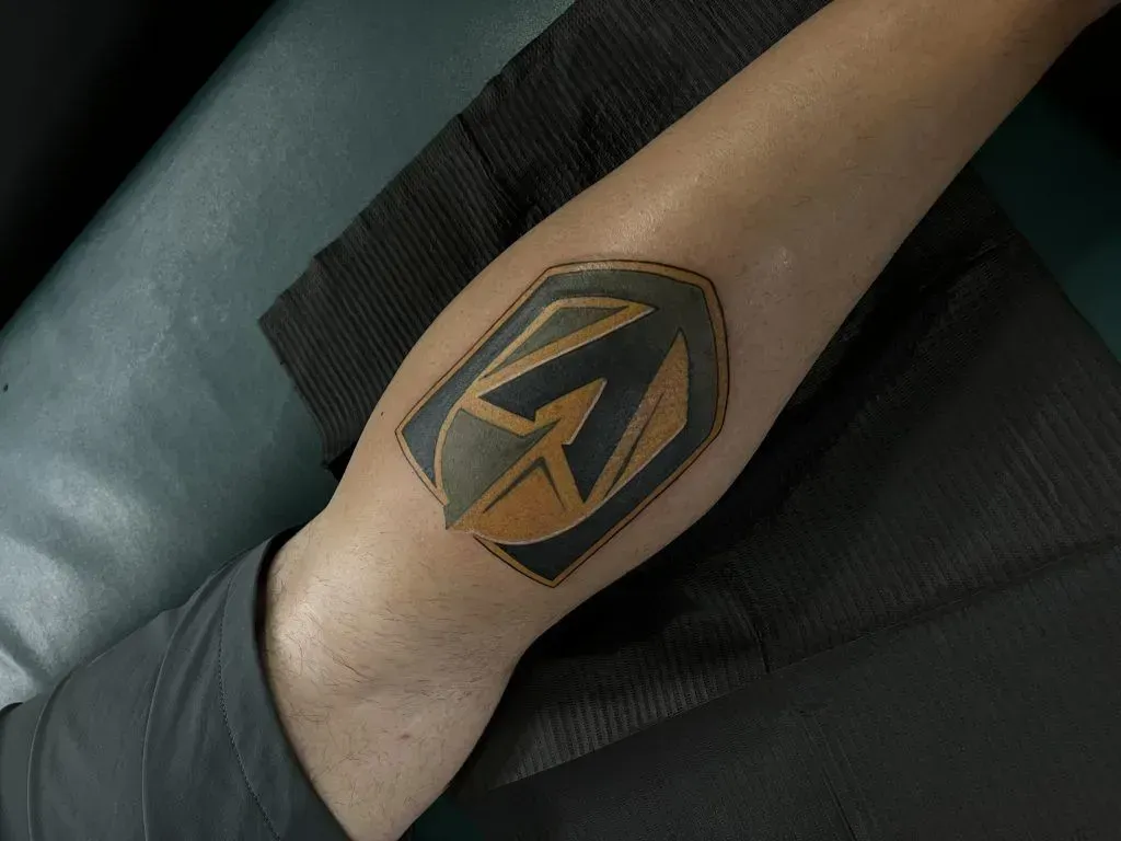 Tattoo of a black and gold logo, possibly sports-related, on a forearm.