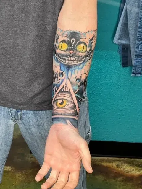 Tattoo of a Cheshire cat above an all-seeing eye on a person's forearm, with blue and yellow coloring.