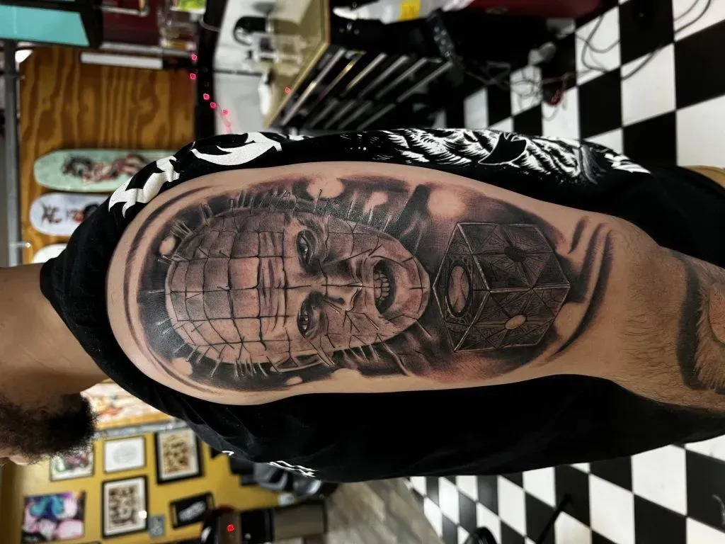 Tattoo of Pinhead from Hellraiser and a puzzle box on a person's upper arm, inside a dark tattoo parlor.
