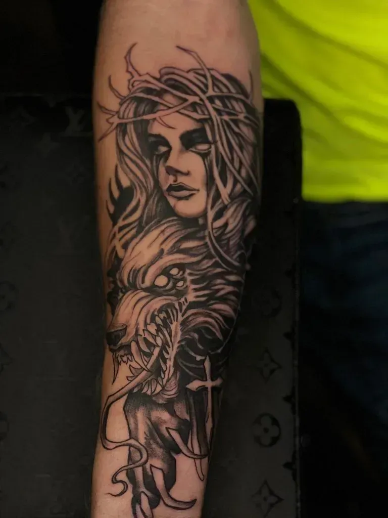 Tattoo on forearm: woman with crown of thorns, wolf with open jaws, and a cross.