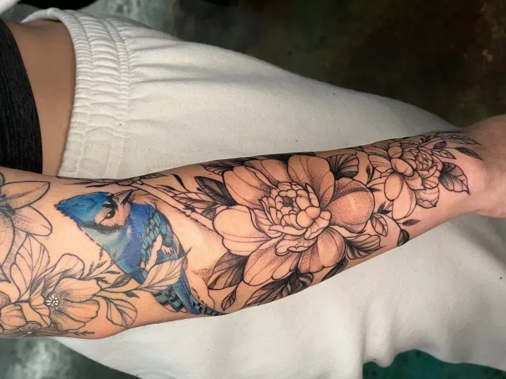 Arm tattoo featuring a blue jay, large flower, and floral elements in black and gray.