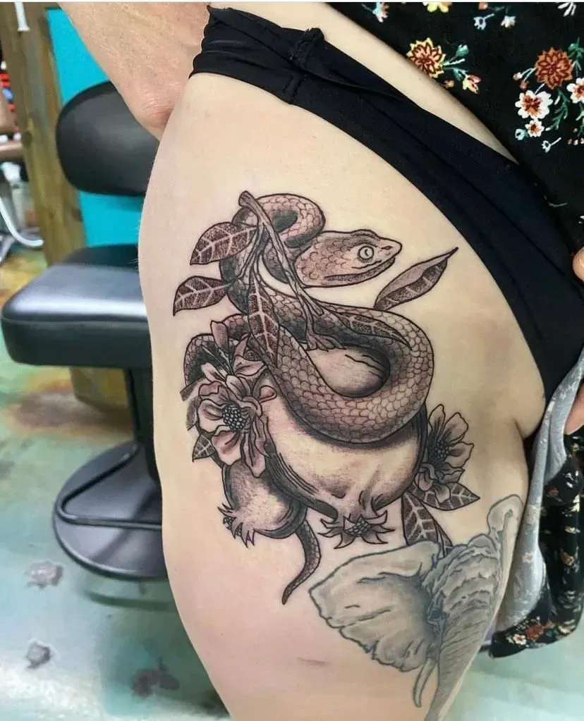 Tattoo on hip of a snake coiled around flowers and a skull. Black and white.