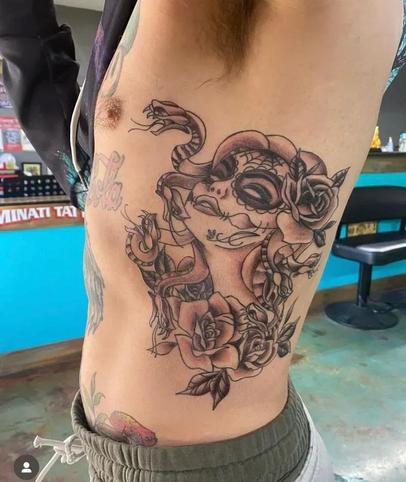 Tattoo on a person's side: Sugar skull, roses, and snake in black and gray. Studio setting.