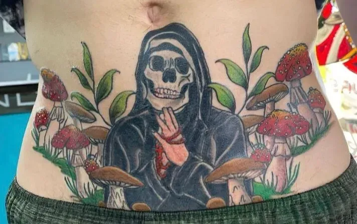 Tattoo on stomach: Grim Reaper in black cloak, surrounded by mushrooms and greenery.