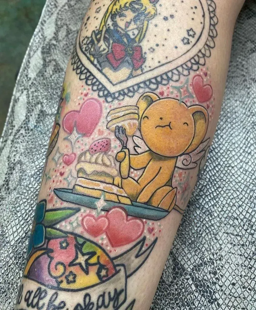 Tattoo of a yellow angel bear eating pancakes, with a Sailor Moon portrait and a “It’ll all be okay” banner.