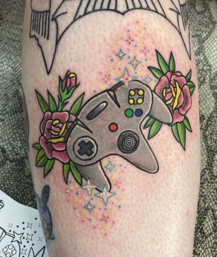 Tattoo of a grey Nintendo 64 controller surrounded by pink roses, green leaves, and stars on skin.