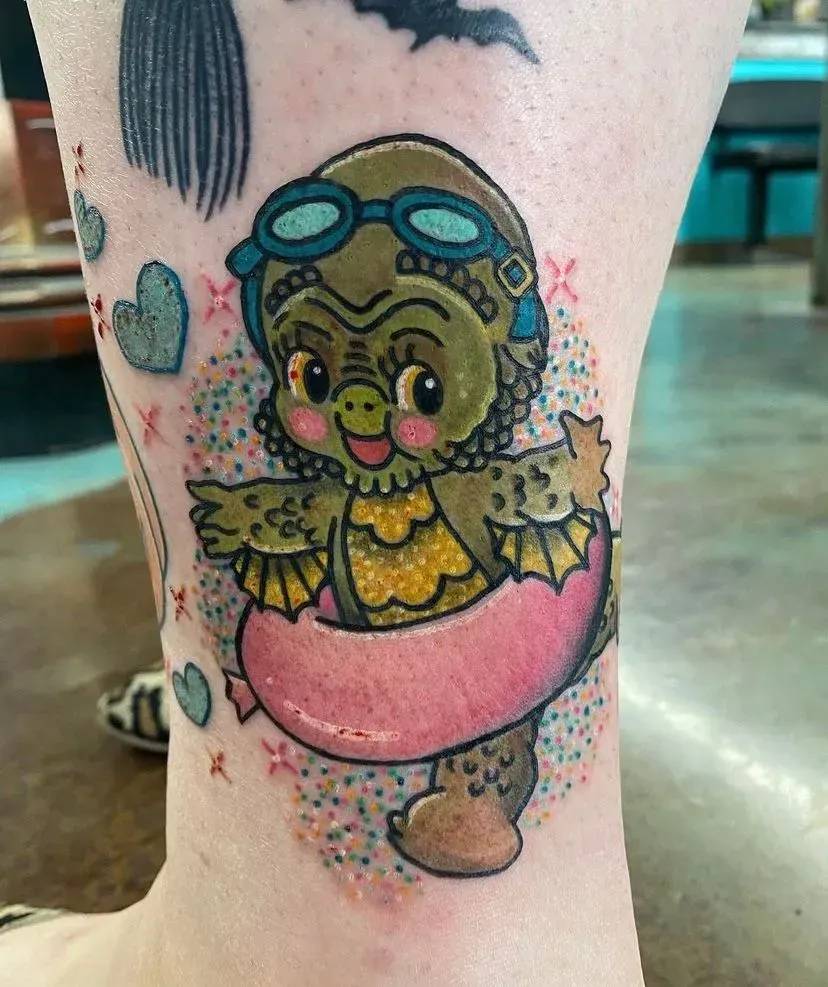 Tattoo of a cute, green Creature from the Black Lagoon wearing goggles and a floatie, surrounded by glittery sparkles.