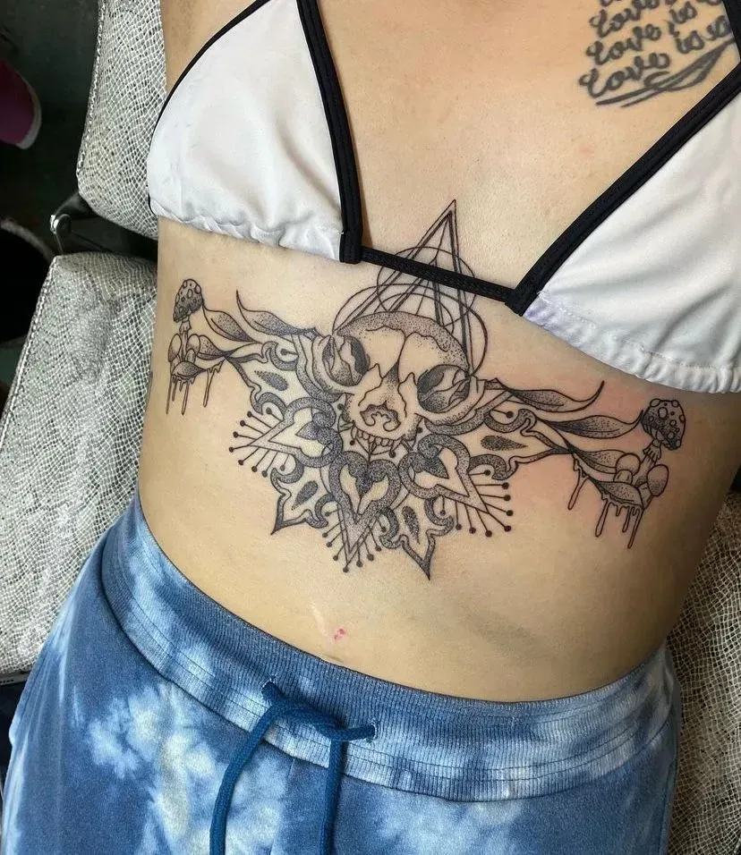 Tattoo on torso: cat skull, geometric design, and mandala elements, white bikini top and tie-dye shorts.