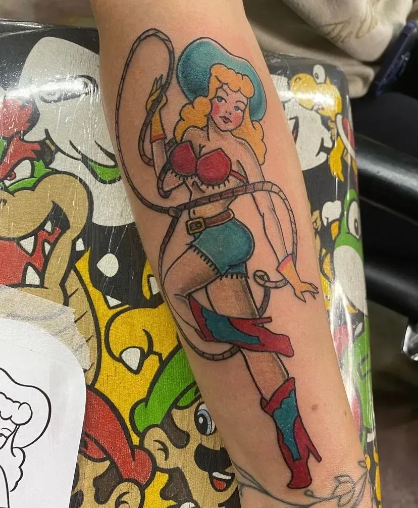 Cowgirl tattoo on arm, holding lasso. Blonde hair, blue hat, red top/boots, turquoise shorts.