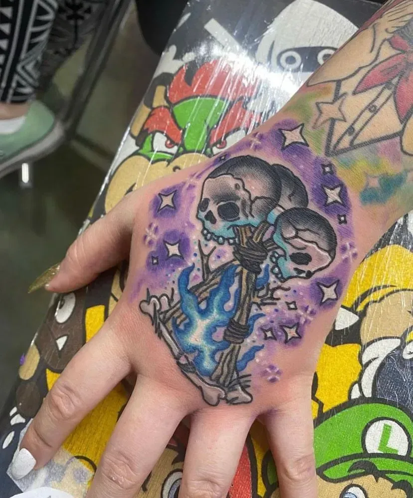 Hand tattoo: three cartoon skulls on bones with blue flames and purple starry background.