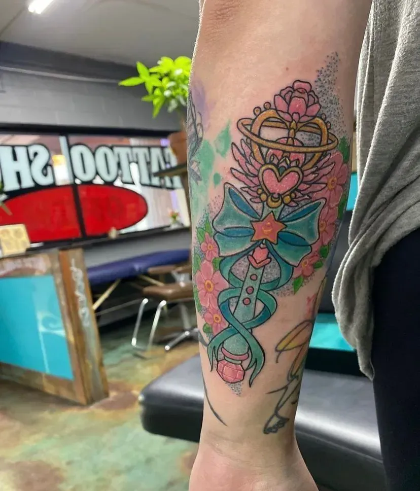 Tattoo of a magical wand with a heart, wings, and bow, surrounded by pink flowers on an arm.